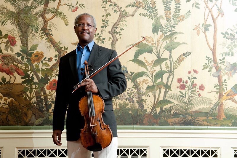 African American violist George Taylor, wearing a robin's egg blue button down shirt, navy suit coat and holding his viola, stands against a colorful wallpapered wall.