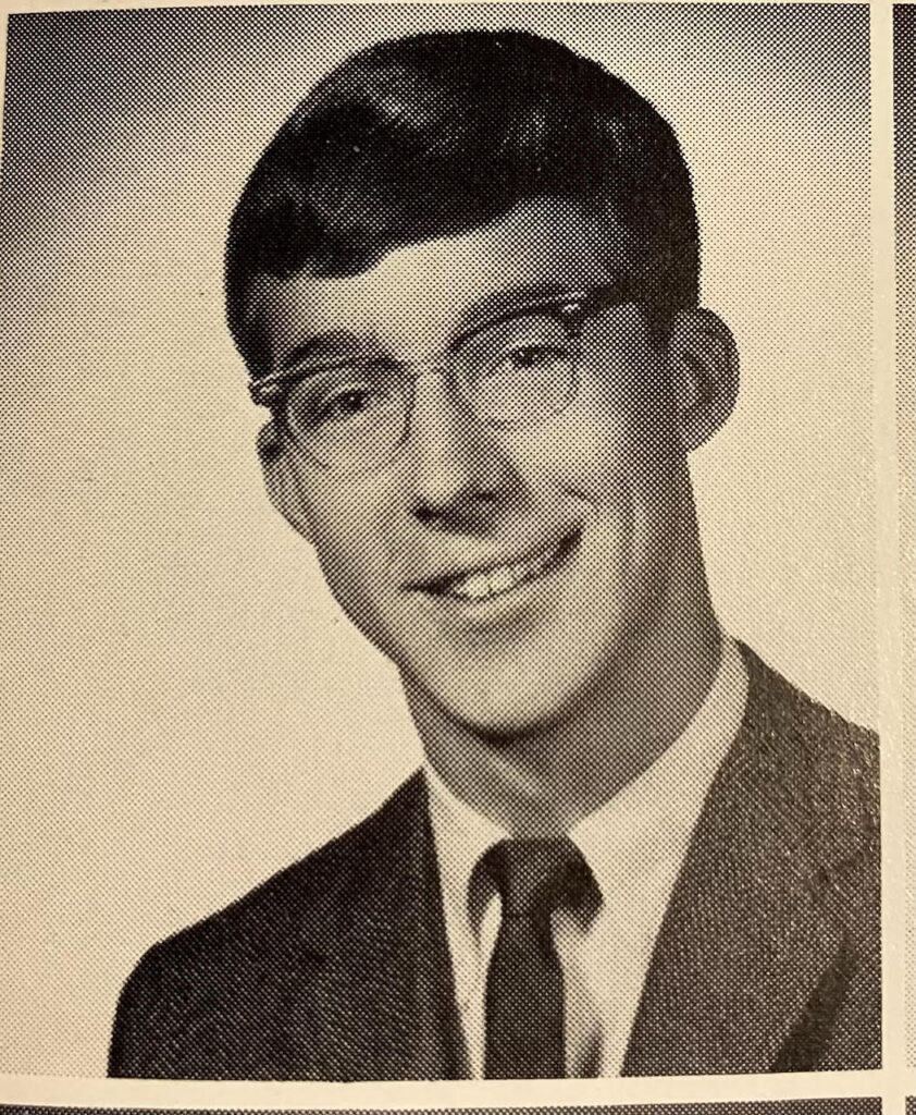 A whie man with dark hair, glasses, wears a white shirt, tie, and suit jacket, smiles in a sepia toned photo