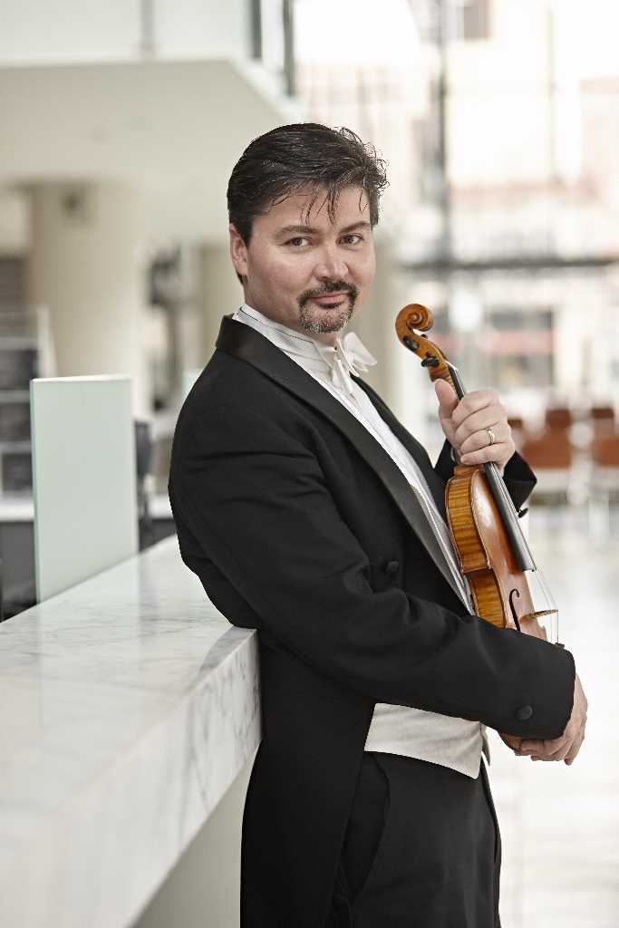 A man wearing white tie and tails holds a violin in both hands, and leans against a railing.