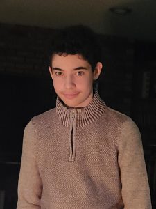 A teenage boy with a closed mouth smile in a tan colored sweater against a black backdrop