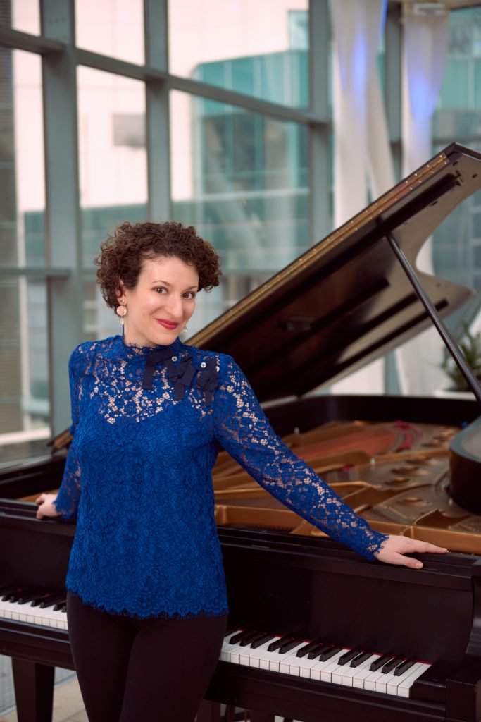 A woman with curly hair wearing a royal blue long sleeved lace top leans against an open black grand piano