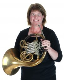 A smiling woman, wearing all black, holds a brass French horn