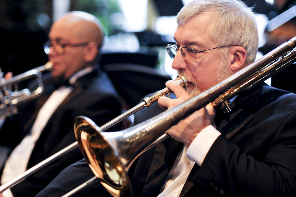 A white man with white hair plays a trombone, a younger Hispanic man plays trumpet in the background