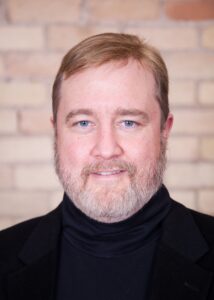 A white man with blue eyes and yellow and gray hair and a beard and mustache, smiles, while wearing a black turtleneck in front of a tan brick wall.