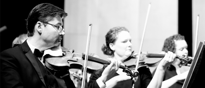 Three violinists in a row, one man with glasses, and two women with curly hair, play