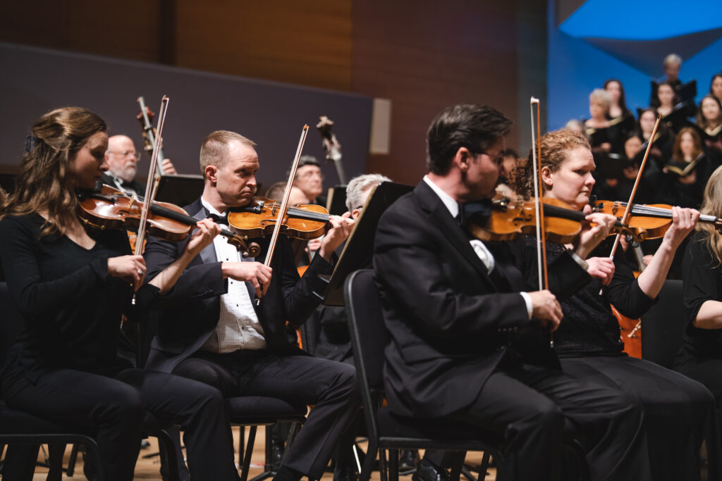 A group of male violinists wearing tuxedos and female violinists wearing black outfits, play together with singers in the background.