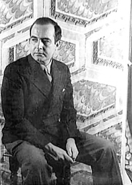 A black and white photo of a middle-aged white man wearing a dark suit and tie, sitting on a chair with a patterned background, looks away from the camera.