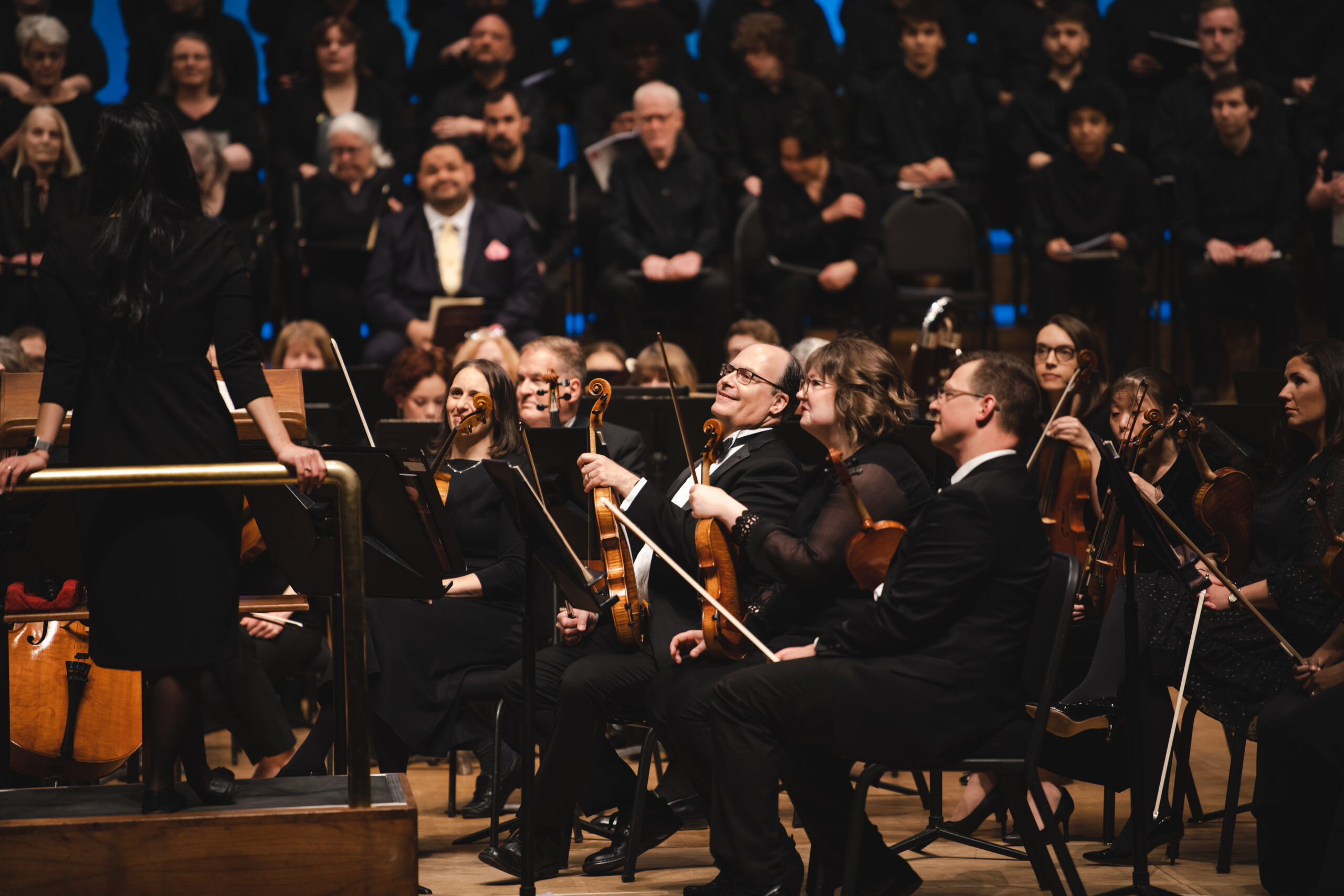 A light colored wooden harp rests on the shoulder of a blond haired woman who we see from the back.