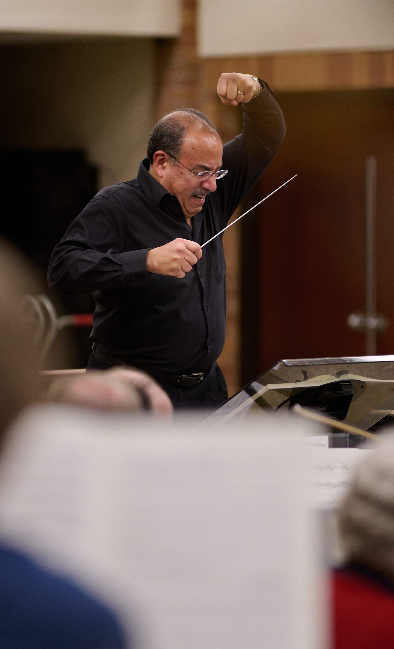 Manny Laureano, wearing a long-sleeve black shirt, holds a baton in his right hand and raises his left fist over his head with an intense look on his face.