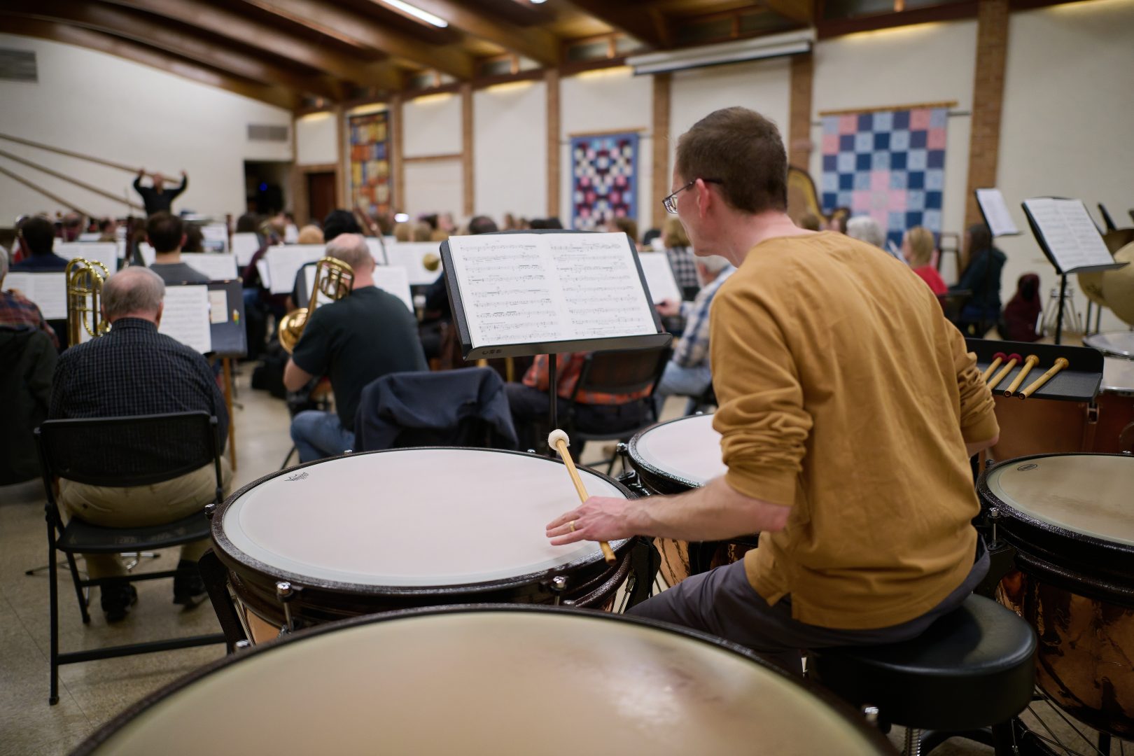 A man in a yellow sweater plays large timpani drums.