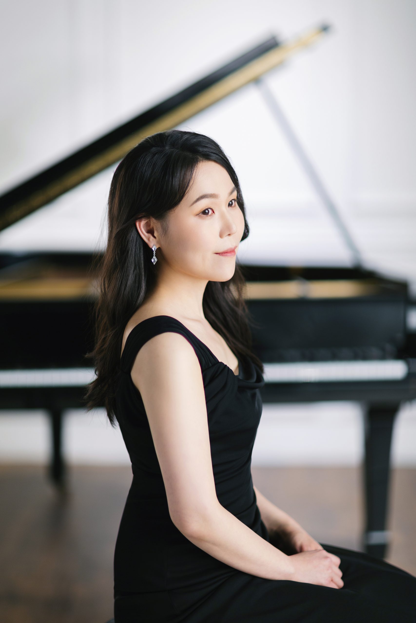 A Korean woman with long black hair and wearing a sleeveless black dress, looks to the right, with an open black grant piano behind her.
