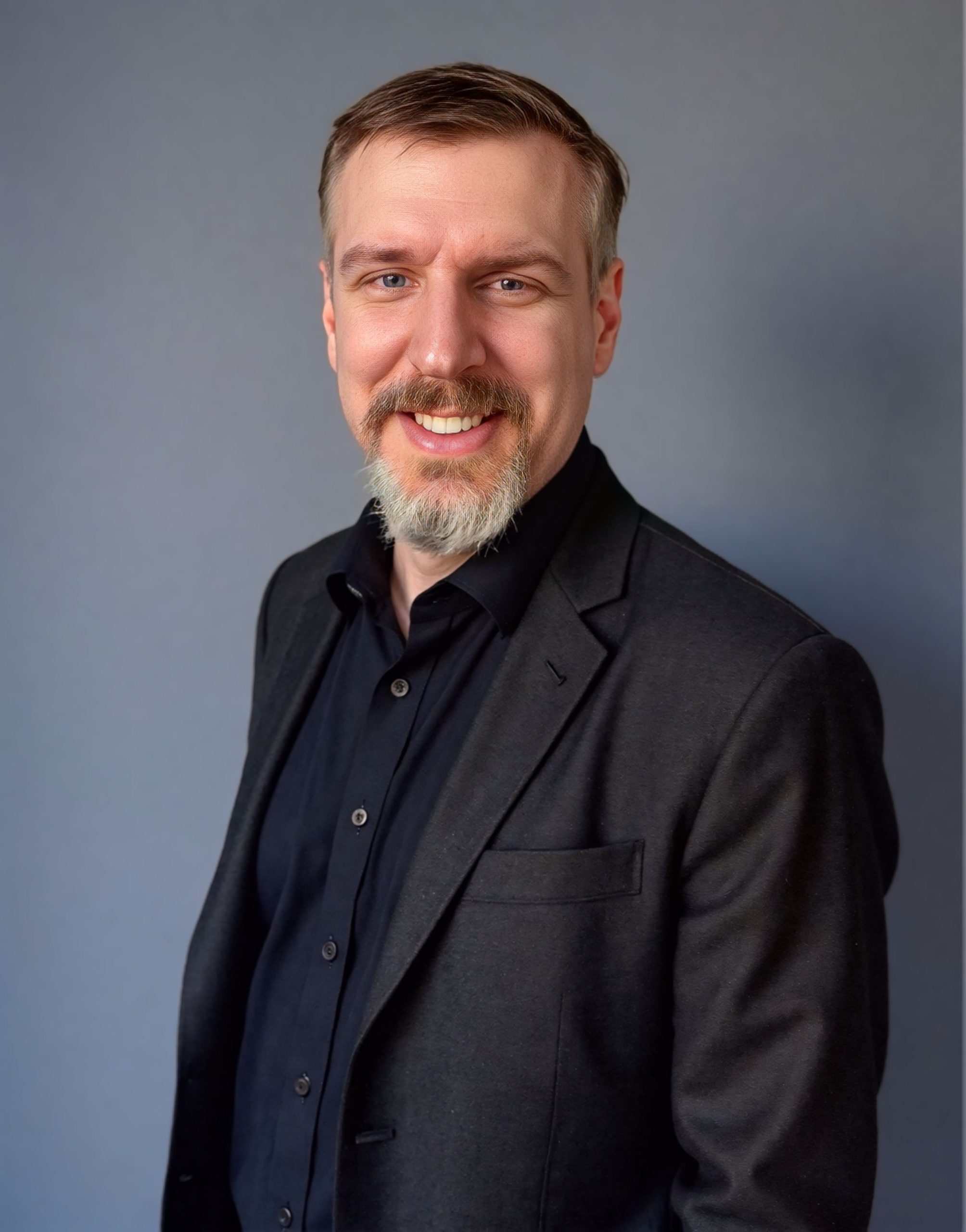 A white man with dark hair and a mustache and goatee, wearing a black shirt and black suit in front of a gray background, smiles at the camera.