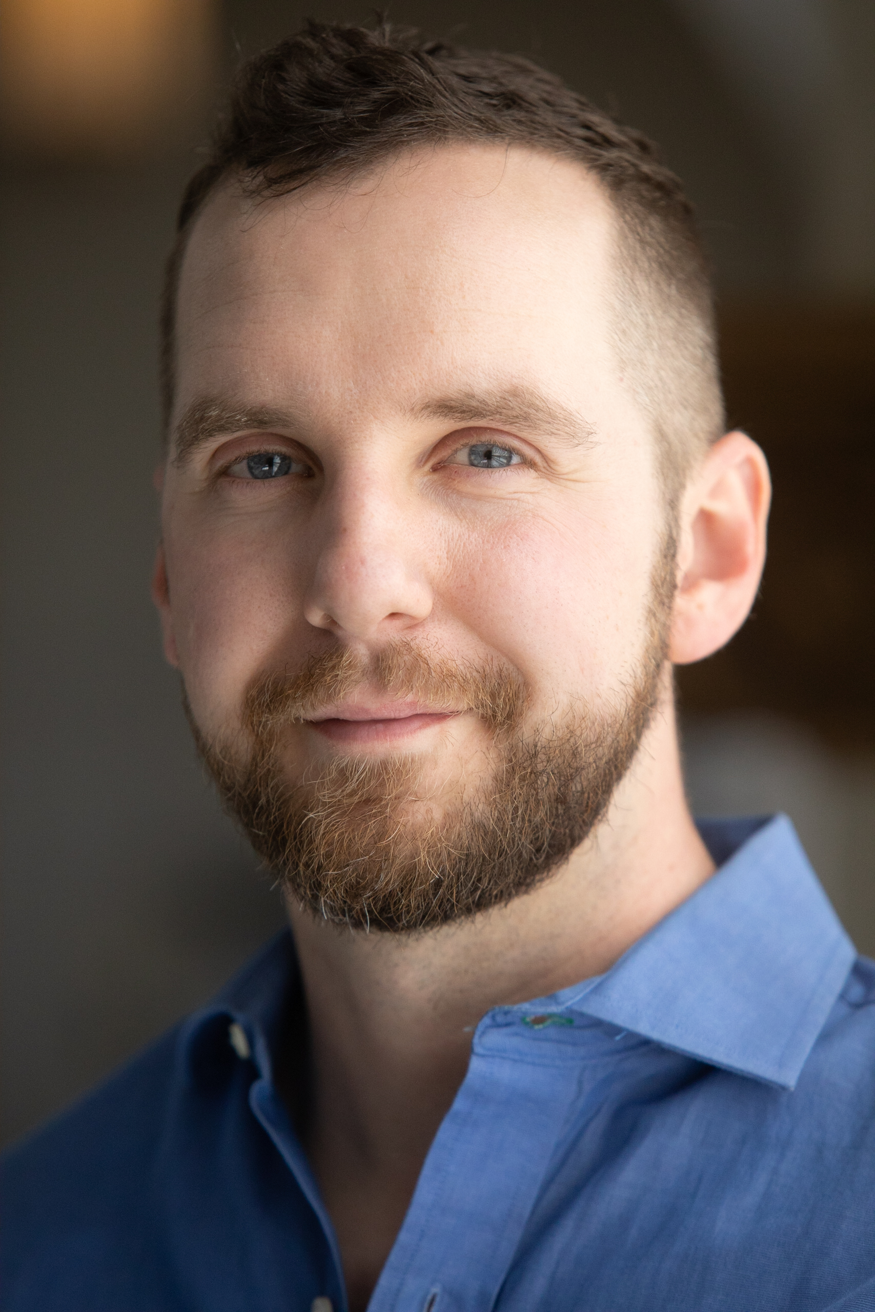 A white man with red-brown hair and a mustache and beard, looks at the camera with a soft smile