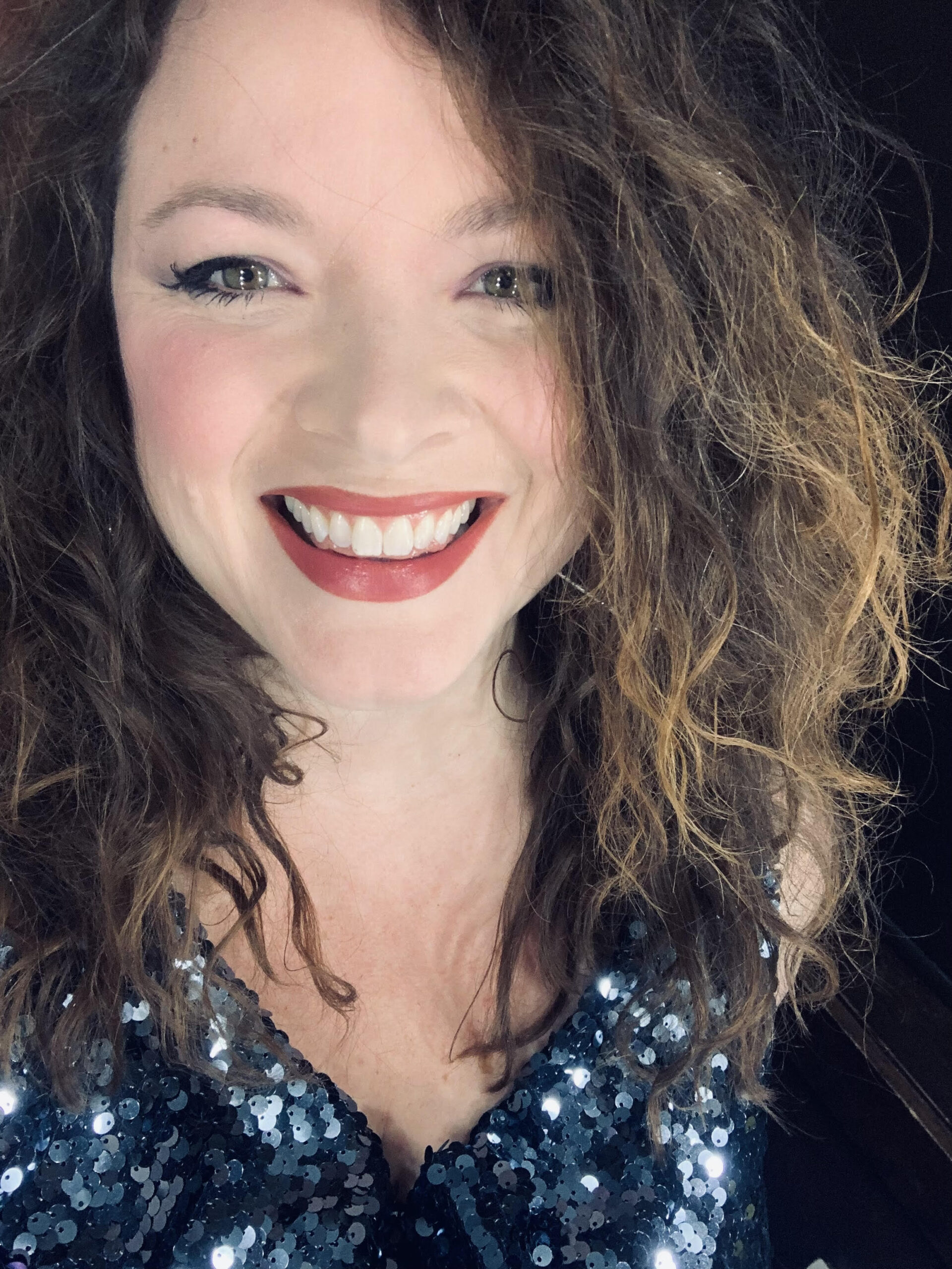 A white woman with brown hair framing her face is wearing a blue sequin top while smiling at the camera
