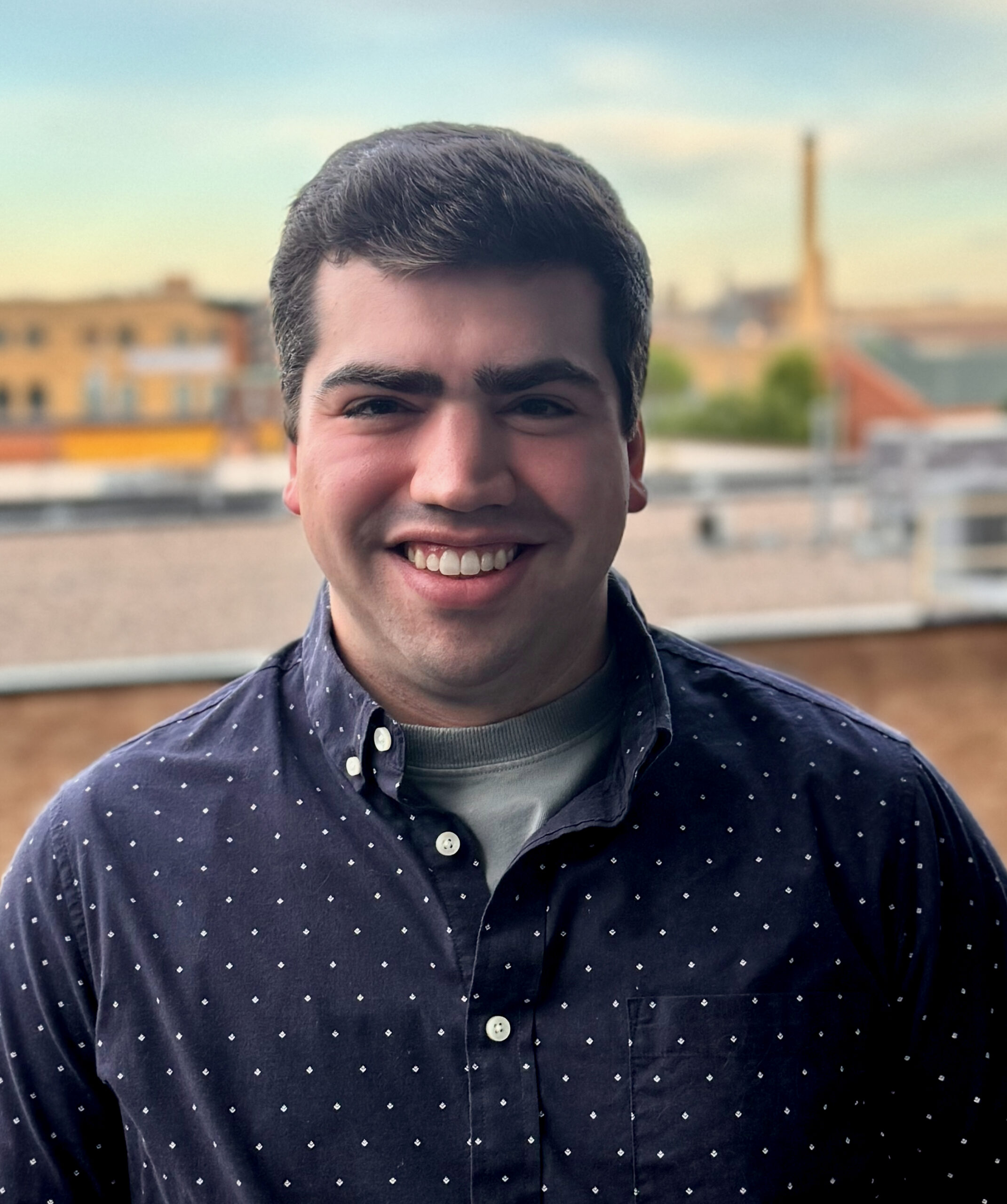 A white man wearing a blue button down shirt, looks directly at the camera with a wide smile