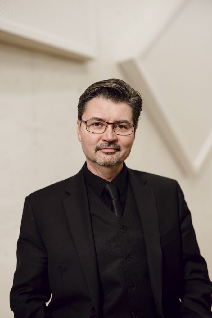 A white man with glasses and facial hair wearing a black suit, shirt, and tie, stands against a white textured background
