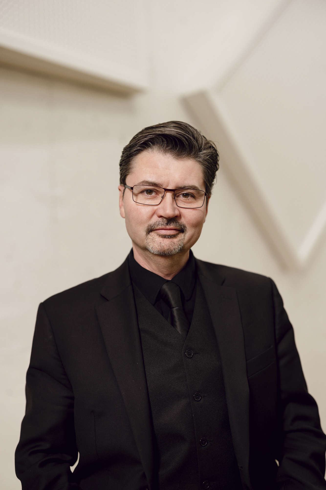 A white man with glasses and facial hair wearing a black suit, shirt, and tie, stands against a white textured background